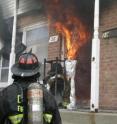 Flames from a living-room pour out the front door of a townhouse in NIST tests that studied conditions that can result in the failure of the facepiece lens of the self-contained breathing apparatus worn by firefighters. Facepieces were outfitted with devices to record temperatures and then mounted on a headform test stand placed outside doorway. The foil-wrapped equipment to the right of test set-up measured thermal heat flux.