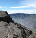 The summit caldera of Tambora, which is about 6 kilometers wide and 7 kilometers long and more than 1 kilometer deep, was created by the 1815 eruption. Gray eruptive products form the top of the caldera wall (foreground) and appear to be 200-year-old pyroclastic flow or late surge deposits. On the floor of the caldera is an ephemeral lake (center) and a small cone from a post-1815 eruption (lower right of lake).