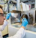 Doctoral canditate Mikko Nisula holds in his hand a sample on a steel substrate. Behind the hand is a ALD reactor.